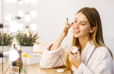 Young beautiful blonde girl cares for face skin with moisturizer in front of mirror.